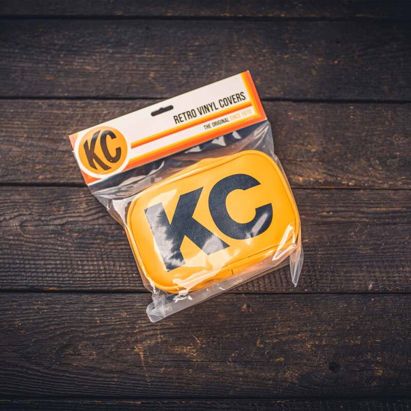 KC HiLiTES Retro 5in x 7in Rectangular Yellow Vinyl Light Cover w/ Black KC Logo (Pair) on wooden background. Main image
